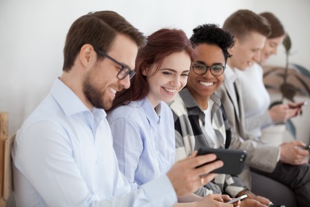 Diverse young businesspeople students friends sitting together in chairs in queue using mobile phones wait job interview or examination. Male showing funny video people laughing, positive emotionsの写真素材