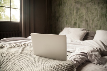 Empty cozy room with an open computer on a bed in bedroom at home no people. Morning light through the window. Weekend and working at home, leisure and lifestyle, wireless technology concept, close upの写真素材