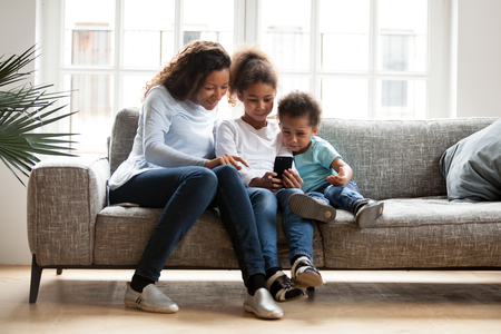 Black family sitting on couch have a fun at home. African mother spend free time with daughter and son using mobile phone taking selfie photo playing new game. Happy family together on weekend conceptの写真素材