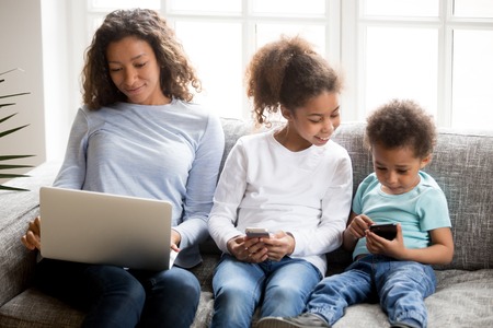 Black family sitting on couch spending free time weekend. African mother using laptop looking at screen working, children have a fun with mobile phones. New generation, addicted with devices conceptの写真素材