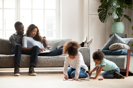 Black family spend free time together in living room at new home. Married couple sitting on couch use computer, little daughter and son have a fun on a warm wooden floor drawing with colourful pencilsの写真素材