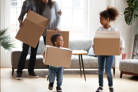 Happy Black African married couple with little kids standing in living room at new home. Small playful adorable kids helps carrying cardboard boxes. New rea estate dwelling, loan and mortgage conceptの写真素材