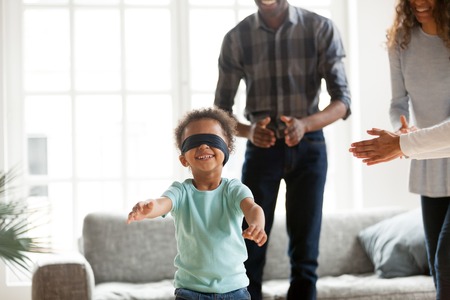 Black african happy family with little children have a fun enjoy spend free time on weekend at new modern home. Playful small son with covered eyes playing blindman game hide and seek in living roomの写真素材