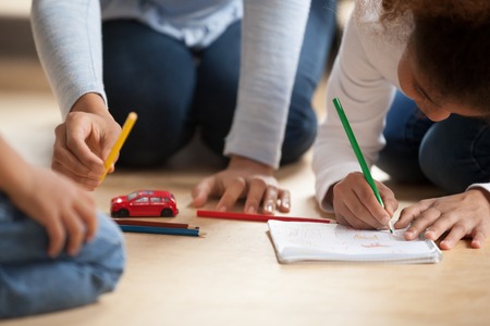 Close up hands of black African mother sitting with toddler son and preschool daughter on a wooden warm floor drawing with colourful pencils spend free time on weekend together in living room at home.の写真素材