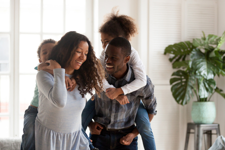 Funny laughing black family hugging with adorable little children have a fun standing in sitting room together at home. Black husband and wife preschool daughter and toddler son piggyback ride indoorsの写真素材