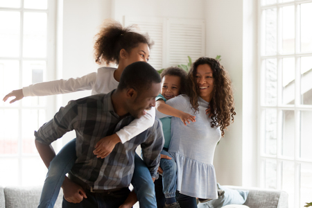Playful laughing black family hugs with adorable little children have a fun standing in sitting room together at home. Black husband and wife preschool daughter and toddler son piggyback ride indoorsの写真素材