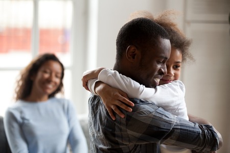 Black African happy family together at home. Close up loving American father strong embrace lovely sweet daughter smiling mother on background sitting on couch. Reunion happy wellbeing family conceptの写真素材