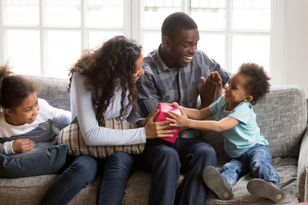 Whole black American family sitting on couch in living room spend time together on weekend at home. Mother helps little adorable son to open pink gift box all family feels happy. Life events conceptの写真素材