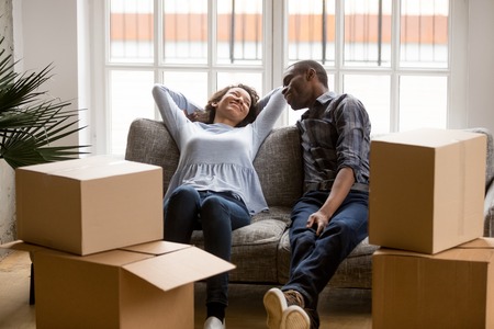 Black married couple sitting on couch in living room at house. Smiling happy wife and husband...