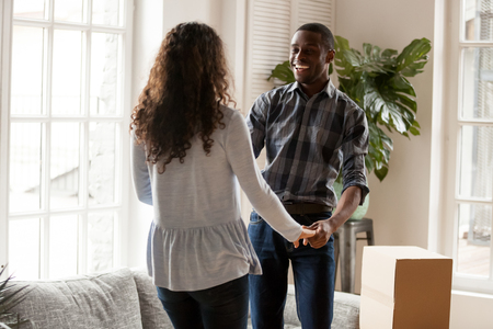 Black couple standing in living room at new house. Smiling husband and wife dancing holding hands celebrate moving at new dwelling feeling happy. Buy real estate property mortgage and relocate conceptの写真素材