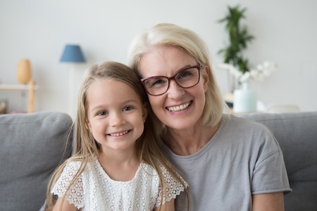 Portrait of smiling cute little girl posing with grandma hugging and cuddling at home, happy...