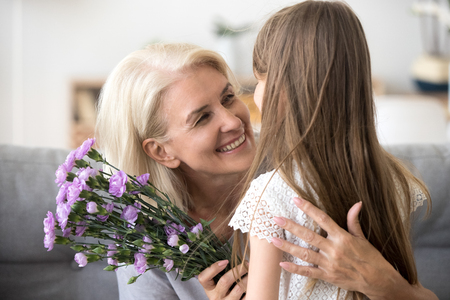 Happy grandmother hugging small cute grandchild thanking for flowers presented, excited granny embrace granddaughter congratulating her with birthday, making surprise presenting bouquetの写真素材