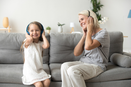 Excited grandmother and smiling little granddaughter listen to music in headphone, grandma and grandchild have fun relaxing on couch at home together, granny enjoy spending time with young generationの写真素材