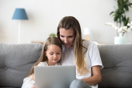 Mother and daughter sit on couch using laptop watching video or cartoon online, cute little girl spend time with mom, relax at home browsing computer or shopping together. Young and technology conceptの写真素材