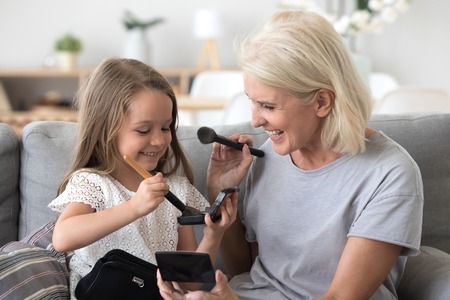 Excited grandmother and granddaughter have fun sitting on couch playing together, happy granny and grandchild do girly things putting make up on faces and laughing, cute girl smiling painting grandmaの写真素材
