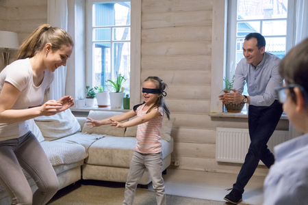 Happy cheerful married couple with small preschool playful children have fun spend free time together on weekend in living room at home blind girl with covered eyes play with family hide and seek game.の写真素材