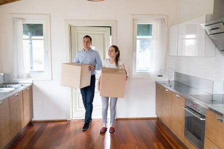 Full length millennial wife and husband hold carton boxes with belongings standing in modern kitchen at home. Attractive happy couple bought new house. Moving relocate day mortgage and tenant conceptの写真素材