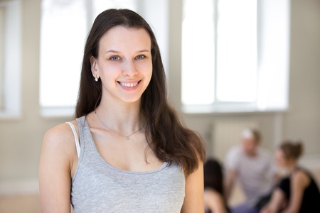 Attractive friendly fitness coach instructor standing at training workout studio, sportive group girls and guys on background. Confident millennial yoga teacher team leader smiling looking at cameraの写真素材