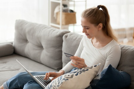 Woman sitting on couch in living room at home alone typing chatting using computer. Positive female holding credit card, buying shopping online paying booking via internet. E-banking service conceptの写真素材