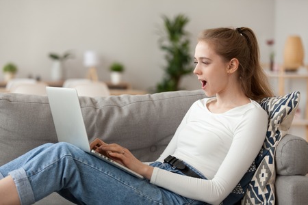 Young positive surprised girl sitting on couch in living room at home alone using laptop. Female looking at computer desktop with opened mouth unexpected great discounts offers, read news on internetの写真素材