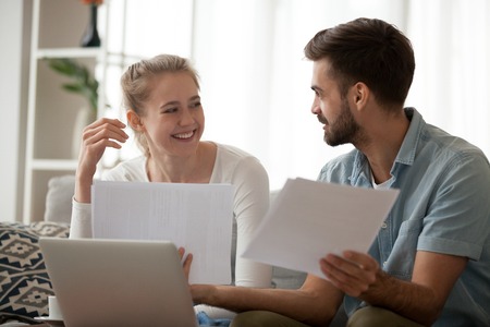 Happy young married couple sitting on couch at home using computer holding paper letter reading received good news about refund or income. Smiling excited wife and husband discussing planning budgetの写真素材