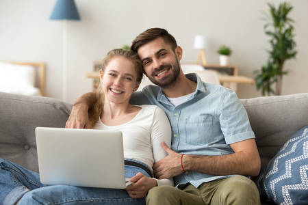 Happy married couple spend time sitting on couch at home. Laughing wife and husband looking at camera. People using computer has a fun watching comedy movie making video call or shopping on internetの写真素材
