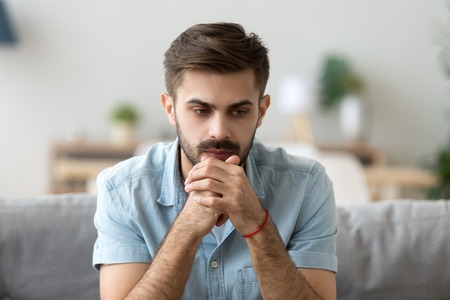 Head shot young millennial serious man sitting on couch home. Portrait of thoughtful pensive...
