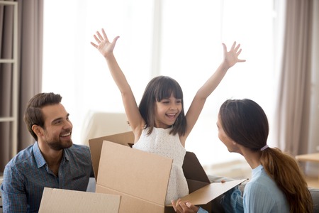 Multi-ethnic family have fun in living room funny daughter jumping out of carton box with hands...