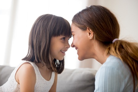 Close up beautiful happy mother and daughter touching with foreheads looking at each other with love tender and devotion, mom expressing support understanding candid emotions sitting on couch at homeの写真素材