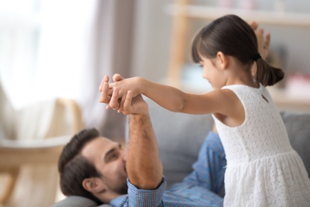 Young father and little daughter play together lying on couch in living room, close up focus on holding hands. Happy family weekend activities best friends warm relations between dad and kid conceptの写真素材