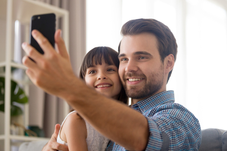 Smiling Caucasian father with little cute mulatto preschool daughter looking at smartphone screen taking making selfie portrait photography sitting on couch. Diverse family having fun together at homeの写真素材