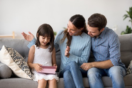Diverse positive smiling family sitting together on couch adorable girl receive from parents mother and father surprise pink gift box kid feels happy and satisfied. Birthday special occasions conceptの写真素材