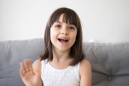 Small girl sitting on couch alone at home. Head shot portrait little adorable daughter waving hand...