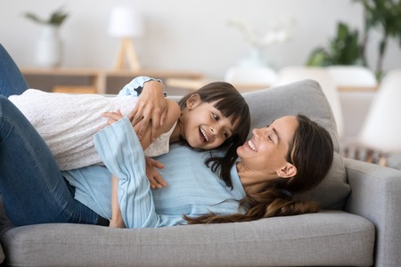 Happy family indoors young cheerful mother or elder sister embrace little cute girl daughter lying on couch in living room at modern home. Kid spending time with loving mom enjoy lazy weekend togetherの写真素材
