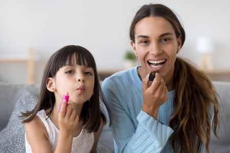 Head shot portrait young beautiful mother with preschool small adorable daughter applying lipstick on lips looking at camera. Little girl copying elder sister have fun together sitting on sofa at homeの写真素材