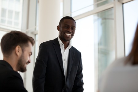 Smiling African American male standing talking during office meeting, happy confident black employee hold business briefing, excited businessman or mentor coaching workers or give trainingの写真素材