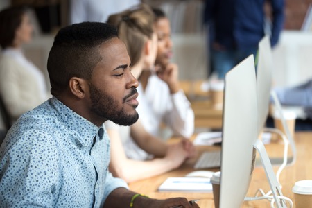 Thoughtful African American male worker looking attentively at computer screen, focused black employee busy working at pc in coworking office, serious afro man study or browsing internetの写真素材