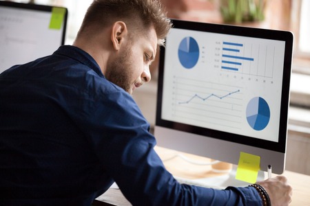 Back view of millennial male employee working on computer in office writing down stats results, focused worker make notes preparing report on pc, serious man busy making research analyzing graphsの写真素材