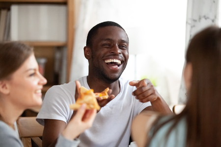 Cheerful african american guy sitting together with caucasian girls laughing spends weekend with best friends. Students millennial diverse people sitting in cafe eating pizza, focus on black maleの写真素材