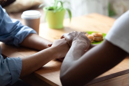 Black african guy holds hand of beloved girl, sitting together at table expressing showing her sincere feelings or apologizing saying sorry. Reliable person, trusted friend, true friendship conceptの写真素材