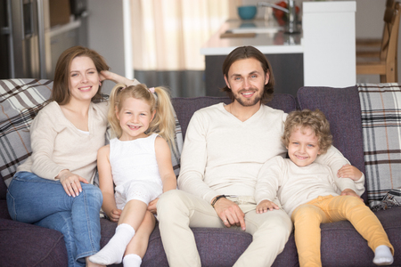Young attractive married couple with little preschool children sitting on couch at new home. Parents and kids smiling and looking at camera. Concept of happy family or buying real estate and mortgageの写真素材