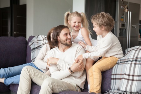 Young attractive married couple with little preschool children sitting on couch in living room at new modern wealthy house. Happy playful parents and joyful kids spending free time or weekend togetherの写真素材