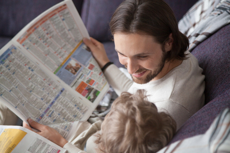 Close up family spending free time weekend at home. Father looking with love tenderness and laughing with little intelligent smart son reading news newspapers sitting together on couch in living roomの写真素材
