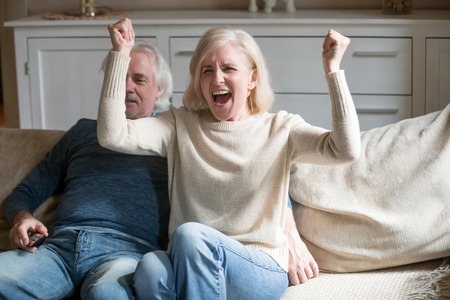 Happy aged couple relax on cozy couch at home watching football game on TV, excited senior wife scream cheering favorite sports team, elderly female supporter or fan rest with husband on sofaの写真素材