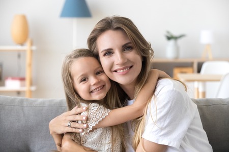 Happy loving young mother hugging little preschool daughter, smiling woman caring mom embracing kid girl sitting on couch at home, mommy and child looking at camera, warm family relations, portraitの写真素材