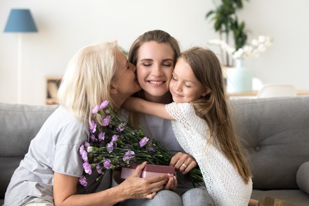 Little kid daughter and old mother kissing embracing young woman congratulating with birthday, senior grandmother and child girl presenting happy mom flowers gift box, 3 generation celebrate togetherの写真素材