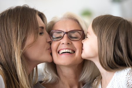 Grown daughter and child granddaughter kissing happy old senior grandmother on cheeks congratulating grandma with mothers day or birthday expressing care and love, three generations of women conceptの写真素材