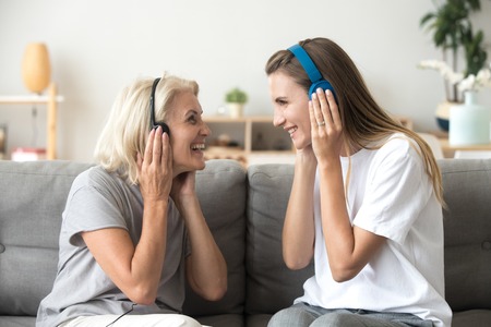 Happy senior mother and adult daughter laughing listening to music in wireless earphones together, smiling young old women wearing headphones having fun enjoy favorite song relaxing at homeの写真素材