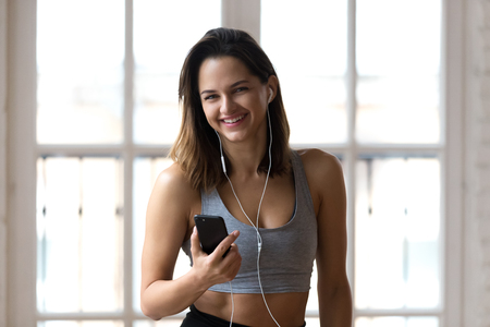 Laughing young yoga instructor experienced trainer resting between sessions listening music using smartphone and earphones in gym. Attractive sporty girl ready for workout smiling looking at cameraの写真素材