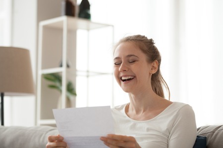 Excited girl sit on couch reading letter knowing good news, happy young female look through paperwork receive positive feedback, smiling woman get document or paper with affirmative responseの写真素材